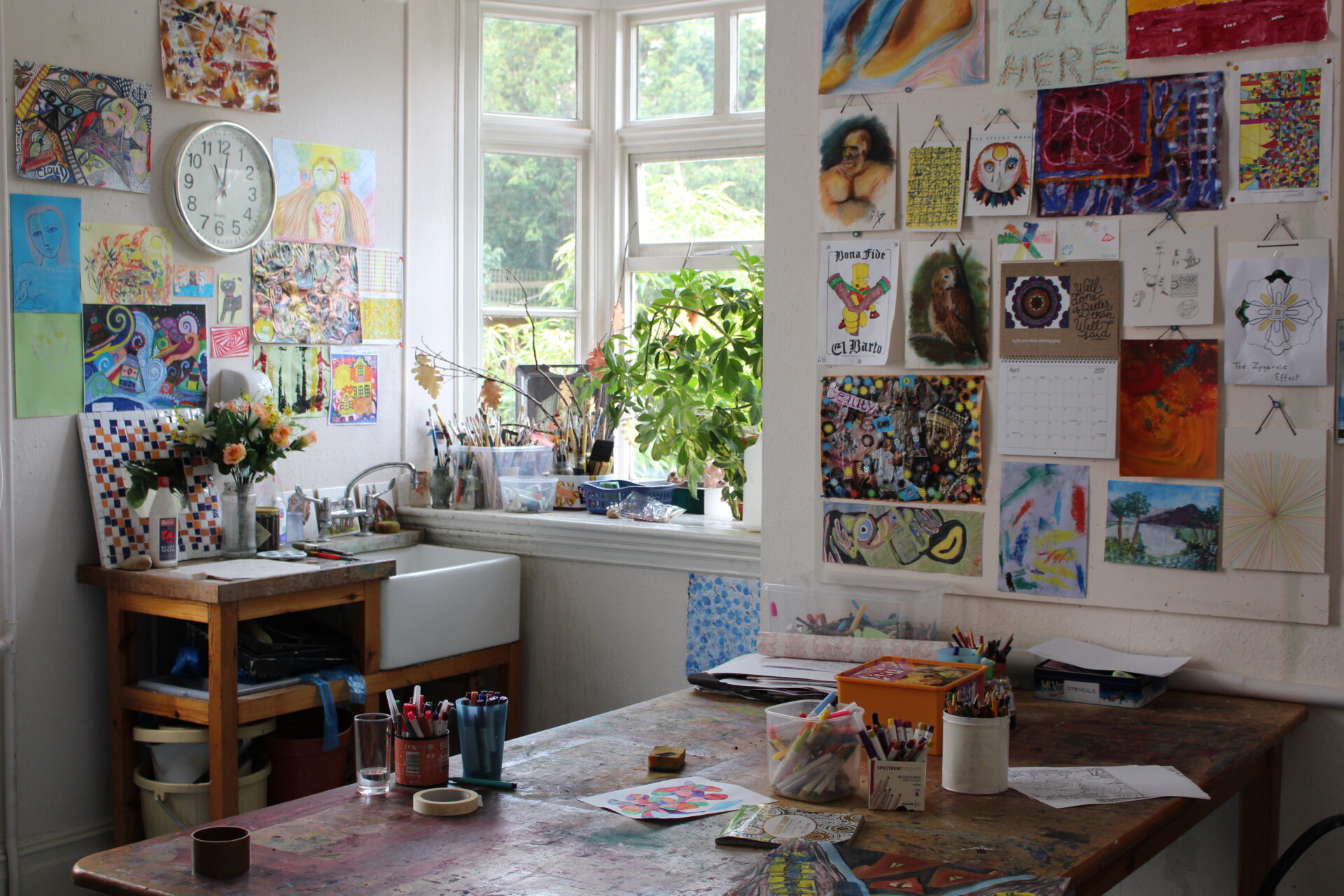 A bright, inviting art room with colourful drawings on the walls, a well-used table filled with pens, pencils and artworks, and leafy plants on the windowsill – a creative space that radiates warmth and imagination.
