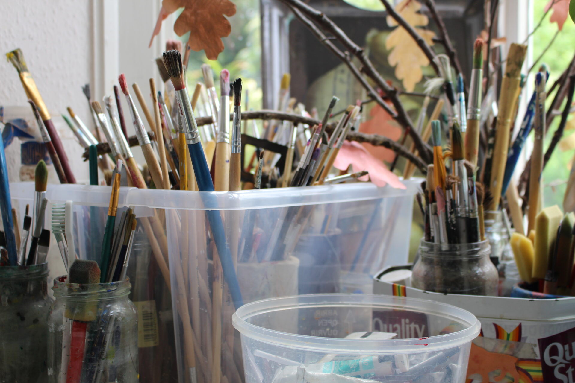 Close-up of jam jars and containers filled with well-used paintbrushes and art tools, set near a bright window with autumn leaves and twigs adding a creative, natural touch – a vibrant symbol of ongoing artistic expression.