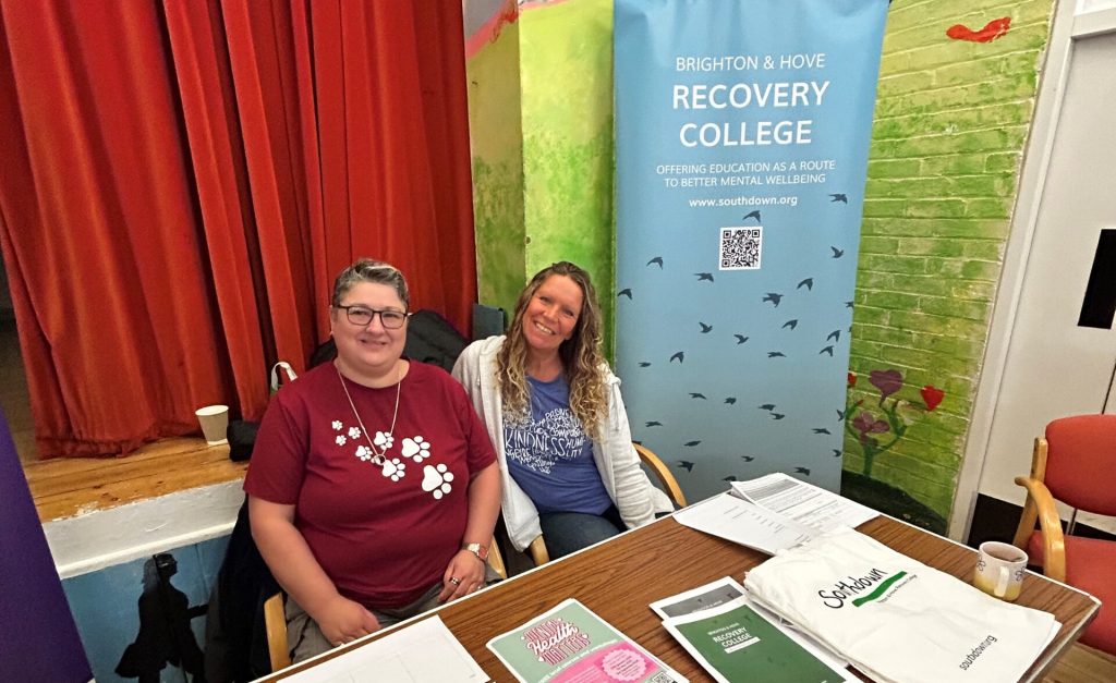Two women seated at a table with informational materials for the Brighton & Hove Recovery College. Behind them is a banner that reads "Brighton & Hove Recovery College - Offering education as a route to better mental wellbeing" with the website "www.southdown.org" and a QR code. The background features a red curtain and a painted wall with a nature theme.||