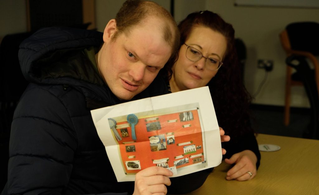 A man in a navy padded jacket sits at a table holding up a colourful poster with photos and blue rosettes. He leans slightly forward and looks directly at the camera. Next to him