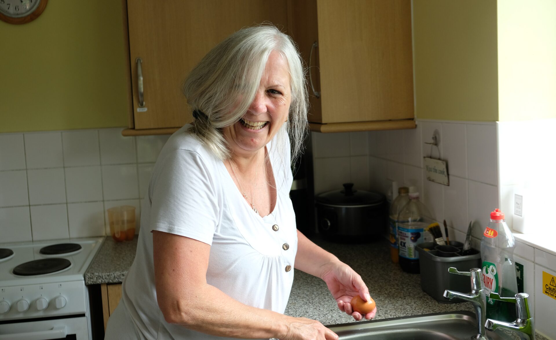 A woman with shoulder-length grey hair and light skin is standing in a kitchen