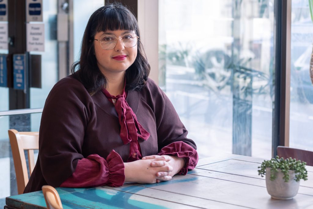 A staff member is sitting down at a table near a window and is looking at the camera.