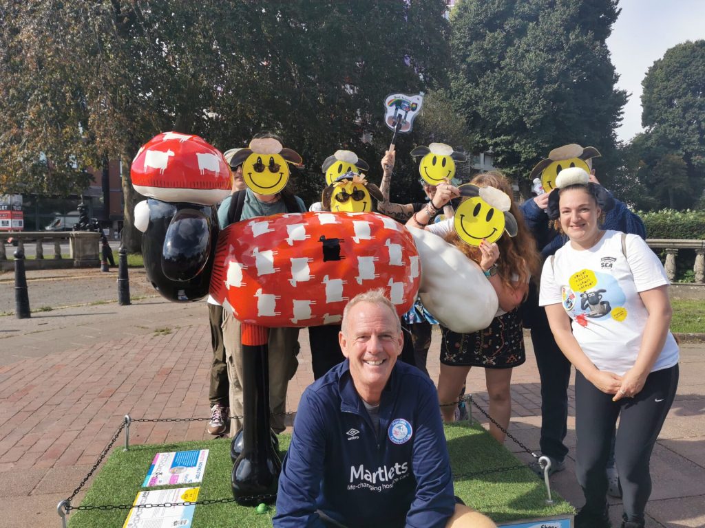 Fatboy Slim is in the foreground. Behind him is a red Shaun the Sheep sculpture and people with smiley face sheep masks|Fatboy Slim is in a park. He is looking directly to camera