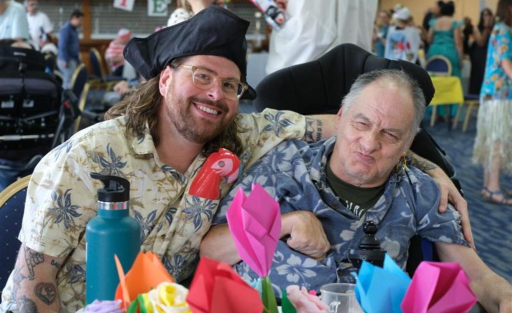 A smiling support worker in a pirate hat poses with a client at the LD Summer Fiesta 2025. Both are wearing tropical shirts and sitting at a decorated table filled with colourful origami flowers. The client