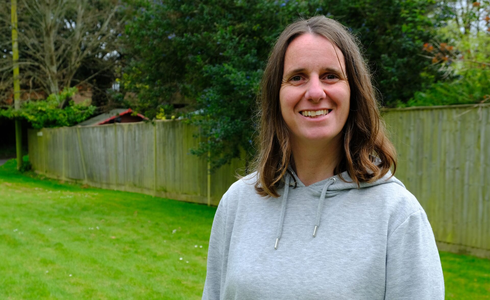 A woman is stood in a garden. She is looking at the camera and is smiling.
