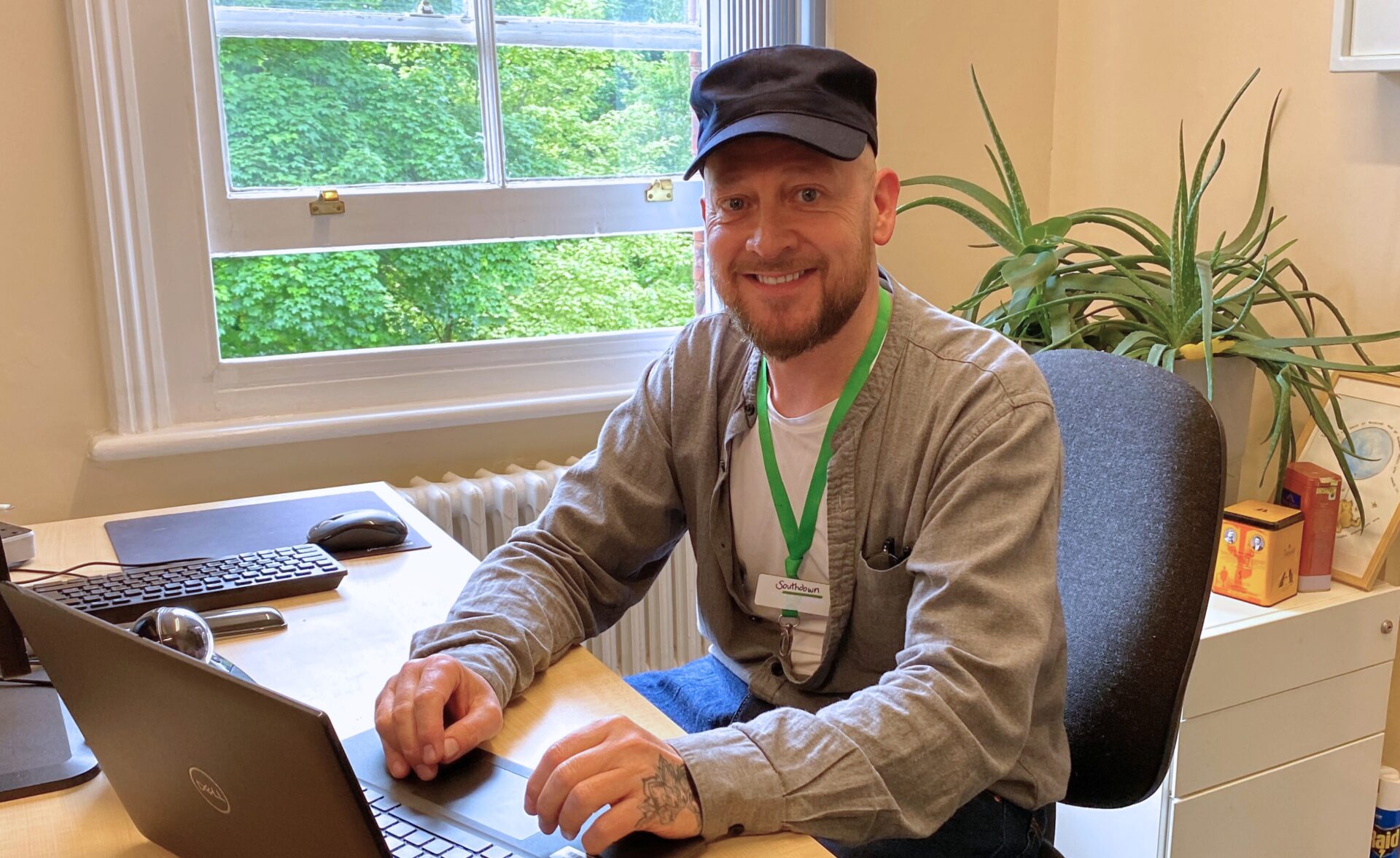 A man is sat at a table inside with a laptop in front of him. Behind him is a window and a plant.