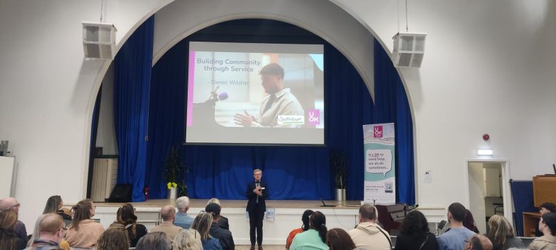 to a seated audience in a community hall. A large screen behind him reads 'Building Community Through Service' and a UOK banner stands nearby
