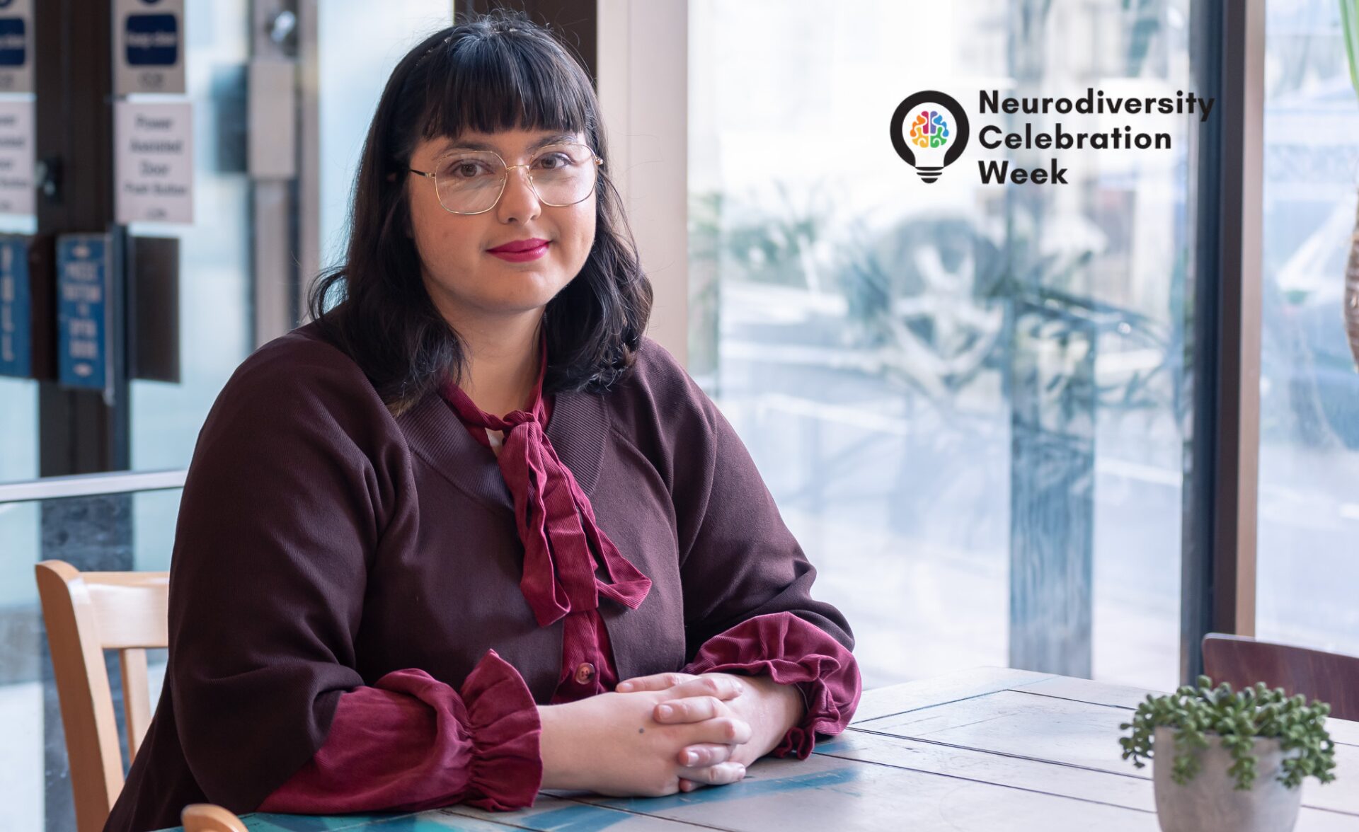 A woman with glasses and shoulder-length dark hair sits at a wooden table in a café. She is wearing a burgundy blouse with ruffled sleeves and a matching bow at the collar. She has a confident and calm expression as she looks directly at the camera. The background features large windows with reflections of the outside environment. In the upper right corner