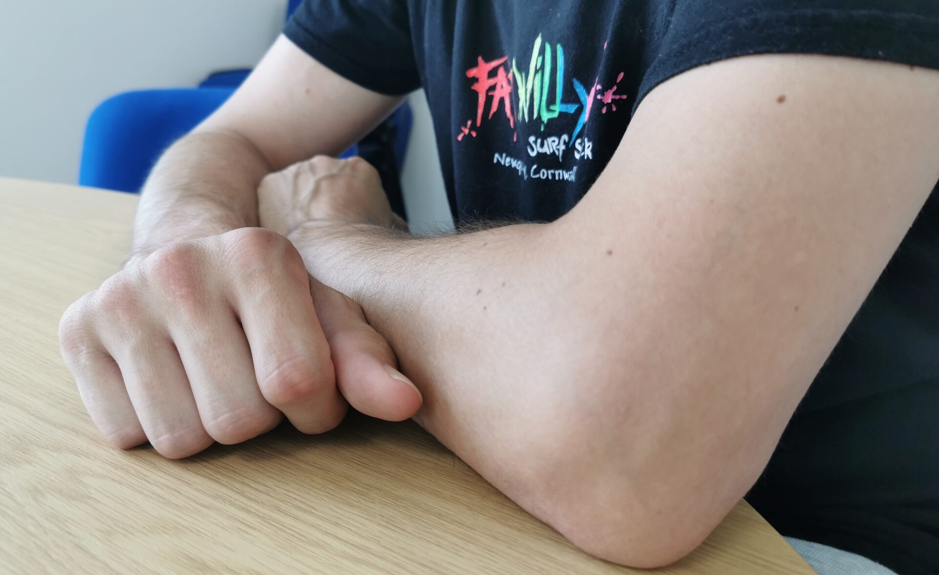 A close-up photograph of a person’s arms resting on a light wooden table. The individual is wearing a black T-shirt with a colourful "Family Surf" logo. The person is a client of the Pathways Home service