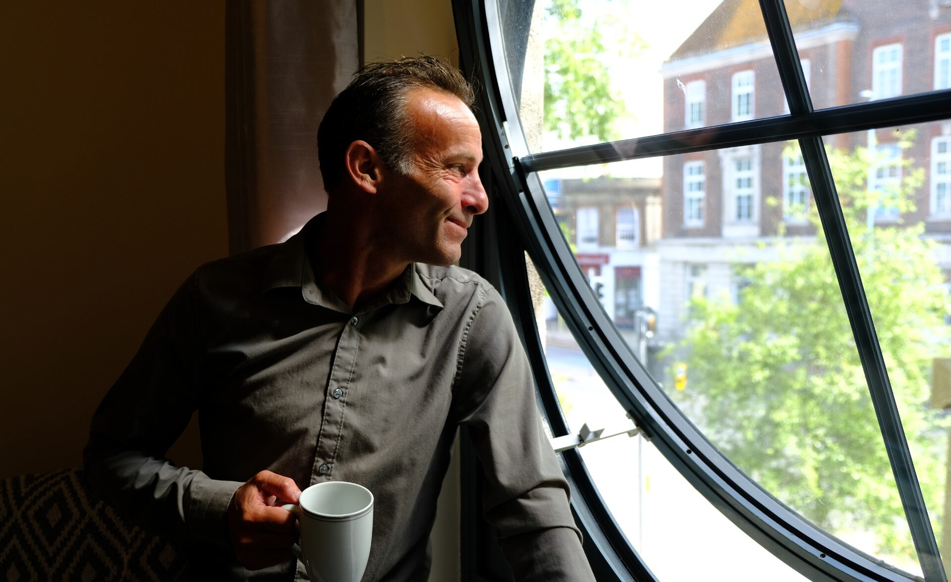 A man wearing a grey shirt holds a white mug and looks out of a large round window
