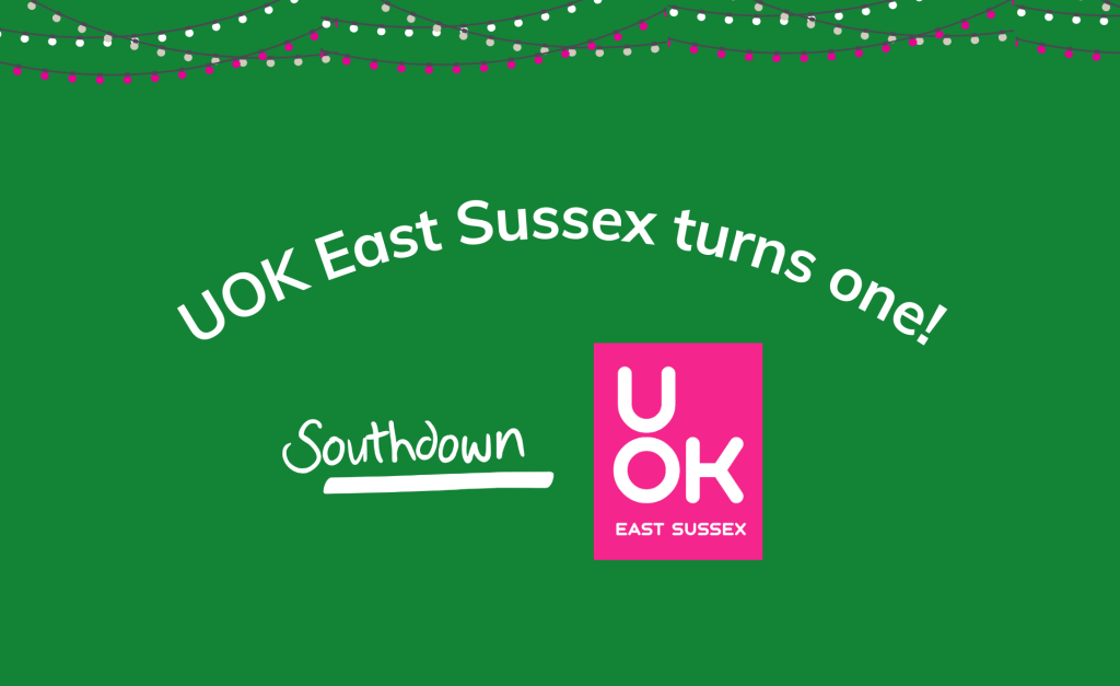 Green background graphic with string lights along the top. Curved text reads 'UOK East Sussex turns one!' Below are the Southdown logo on the left and the pink UOK East Sussex logo on the right.