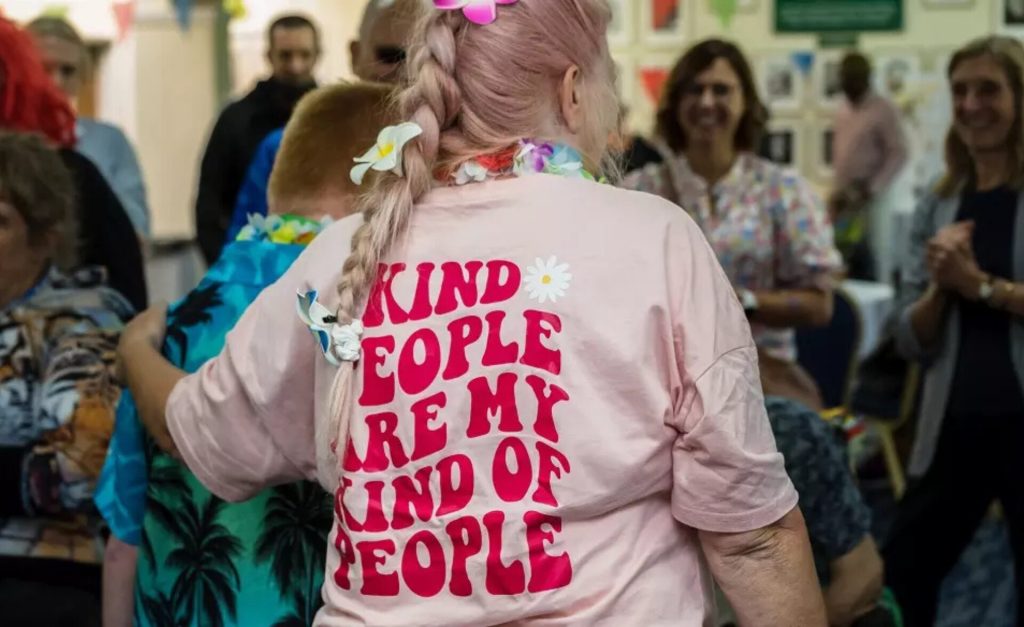 A person with pink hair styled in a braid wearing a pink T-shirt that reads "Kind people are my kind of people" in bold red text with a daisy design. They are participating in a lively Learning Disability Service event.